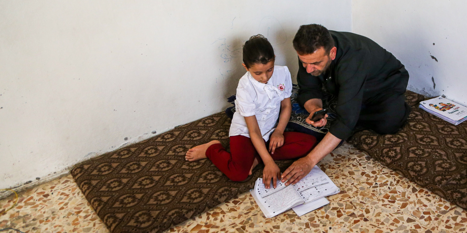 In Northwestern Syria, Rama and her father, Fadi, use his smartphone to watch a virtual Arabic class on WhatsApp, supplemented by textbooks.
