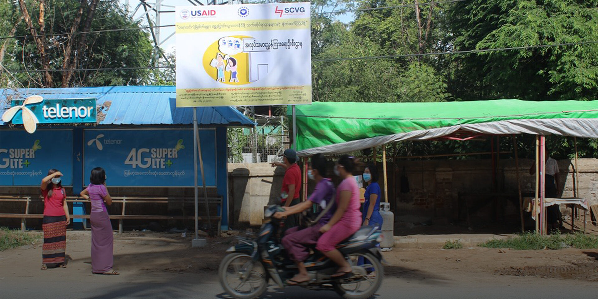 In the Magwe region in September 2020, passengers on a motorbike pass a USAID PRLM billboard about preventing and reporting human trafficking. The Magwe region has a large population of migrant workers, who can have higher risks of becoming victims of trafficking.