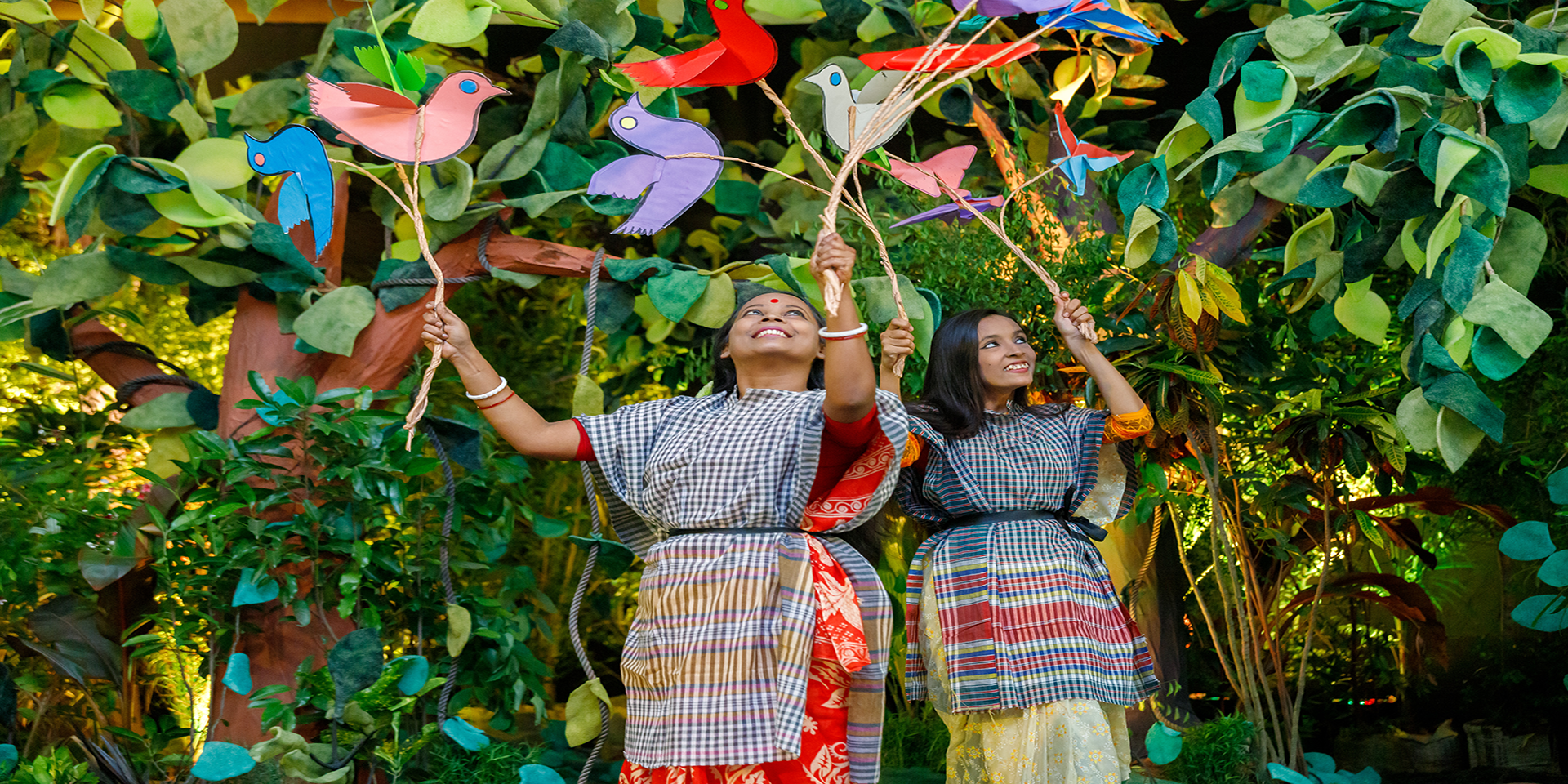 Two young women holding paper birds are part of a performance portraying life inside a forest, in front of the audience attending the launch event of the USAID Ecosystems/Protibesh Activity.