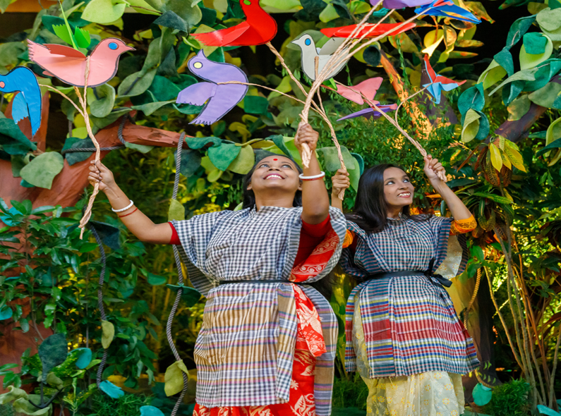 Two young women holding paper birds are part of a performance portraying life inside a forest, in front of the audience attending the launch event of the USAID Ecosystems/Protibesh Activity.