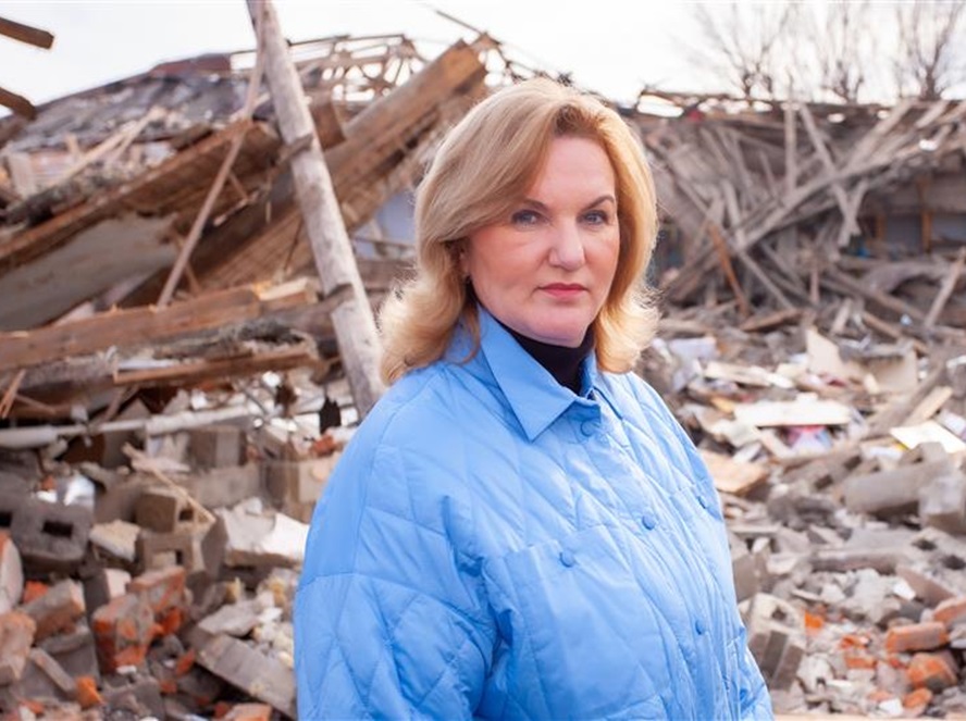 A woman with short blonde hair wearing a blue jacket stands in front of rubble