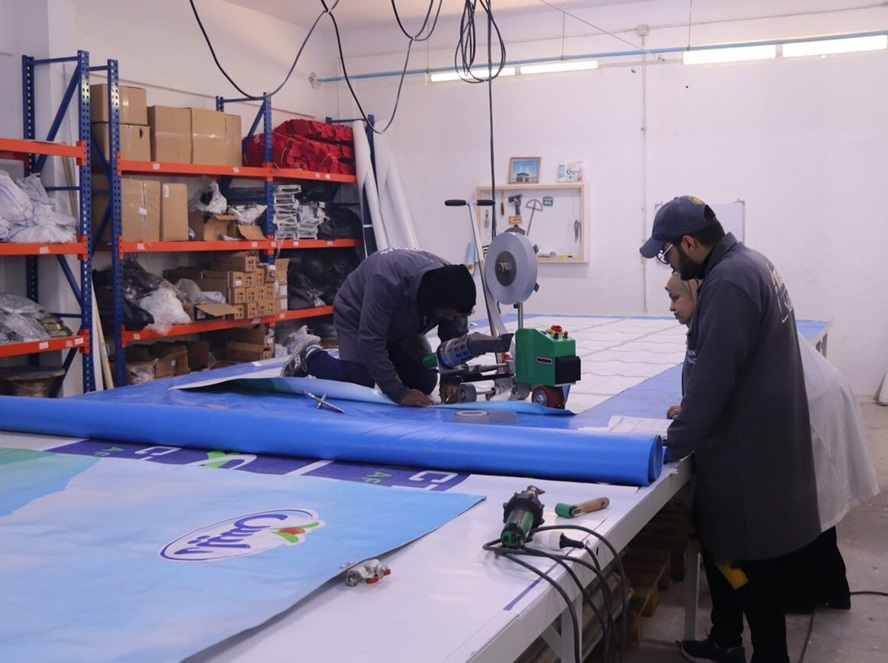 A man sits on a table printing a large banner while two people look on