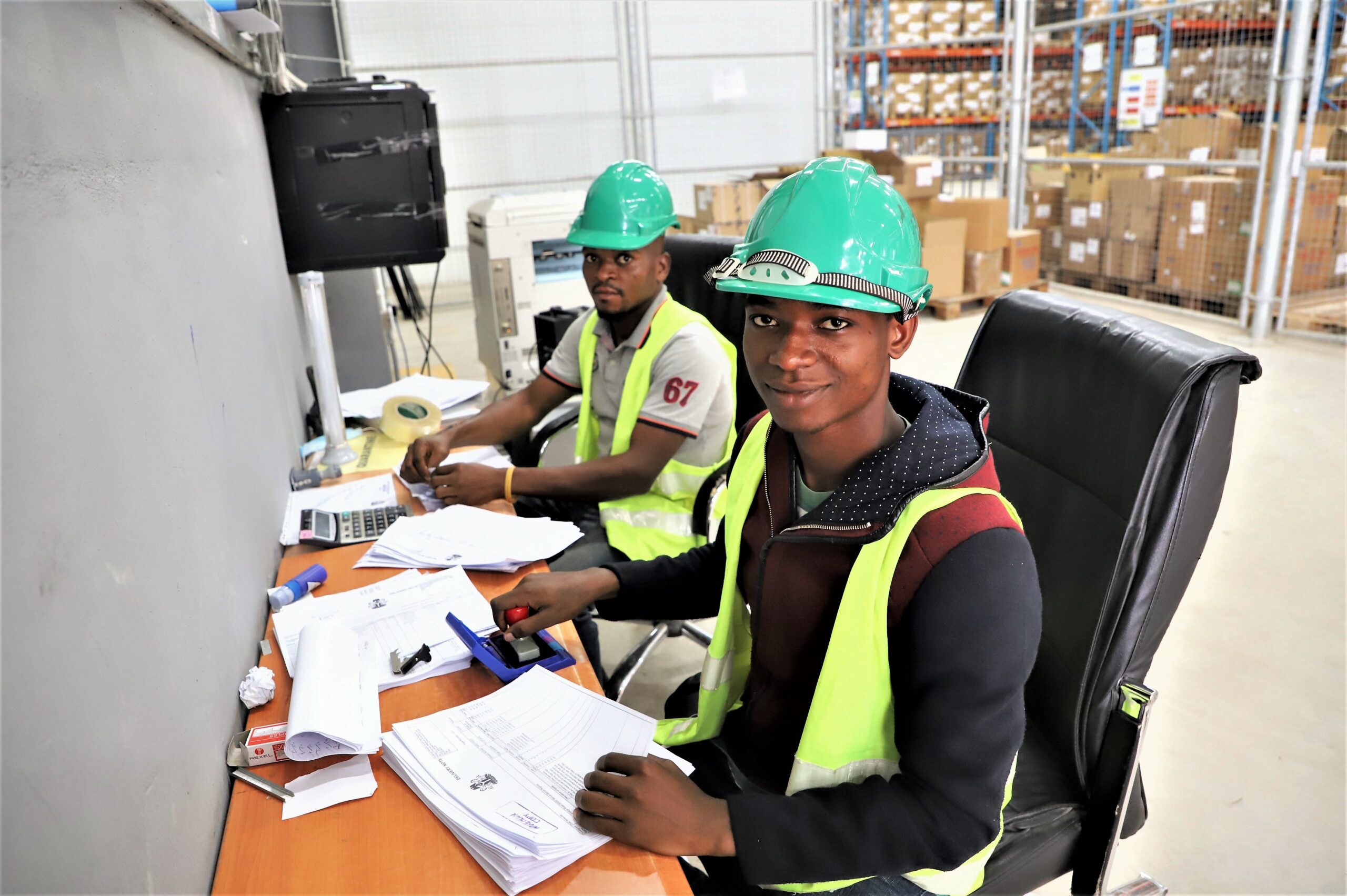 Two men wearing safety gear sit at a desk making copies of documents
