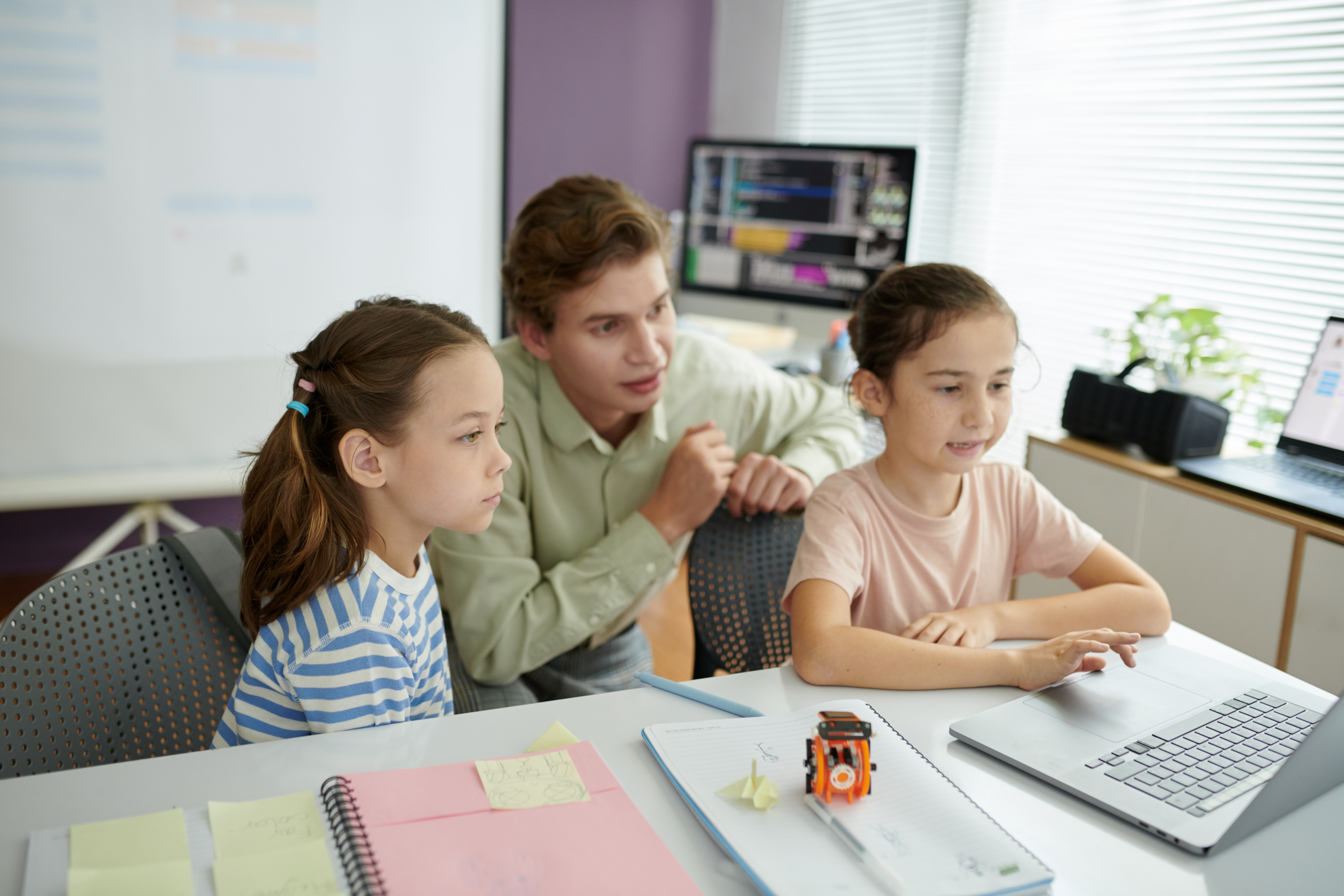 Teacher helping two young children with computer programming project while they are seated at desk. Classroom has educational materials and computer equipment in the background