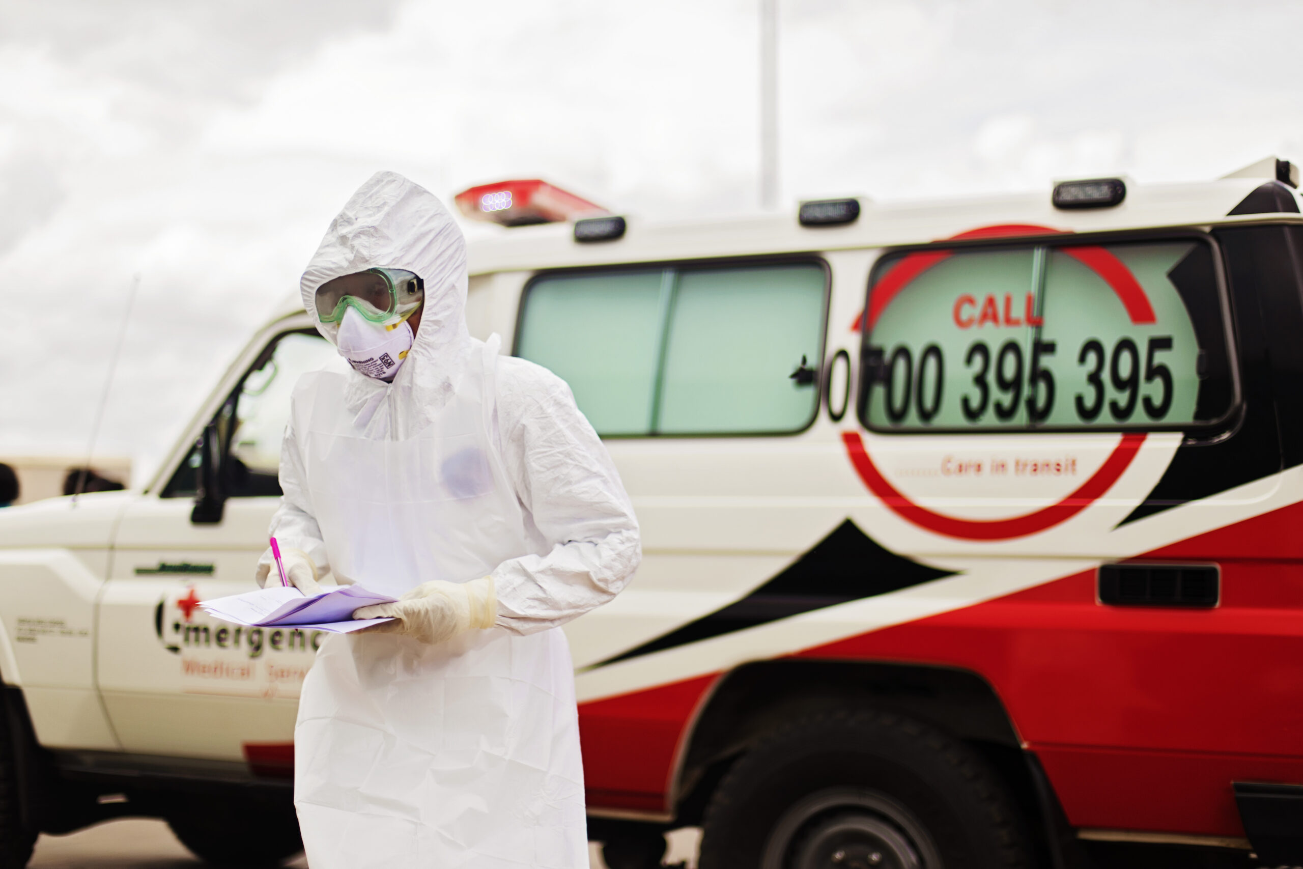 A person in full PPE stands in front of an emergency vehicle holding papers and a pen